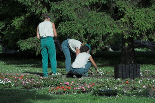 The image depicts three individuals working together in a lush garden with a rich green backdrop of tall, dense trees and a well-maintained flower bed. The garden features a grassy lawn area in the foreground, bordered by a variety of colorful flowering plants arranged in neat flower beds. One person is standing, facing away, wearing a sleeveless top and teal trousers, while the other two are crouching and kneeling, actively tending to the plants; one appears to be planting or weeding, and the other is closely inspecting the soil or flowers. There is a small black garden border or edging visible on the right side of the flower bed, and the scene is illuminated by natural daylight, suggesting a clear weather day. This setting illustrates outdoor garden maintenance, with a focus on flower bed care, and reflects typical landscaping activity in a residential garden in Upminster, suitable for professional gardening services such as those offered by Gardening Services Upminster.